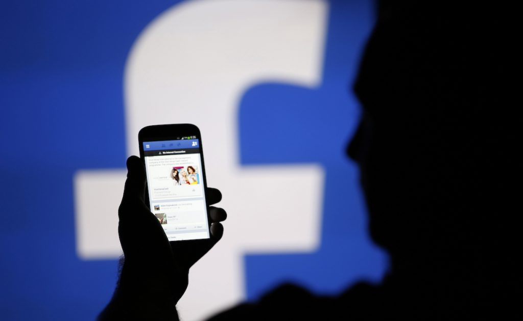 A man is silhouetted against a video screen with an Facebook logo as he poses with an Samsung S4 smartphone in this photo illustration taken in the central Bosnian town of Zenica, August 14, 2013. REUTERS/Dado Ruvic/File Photo