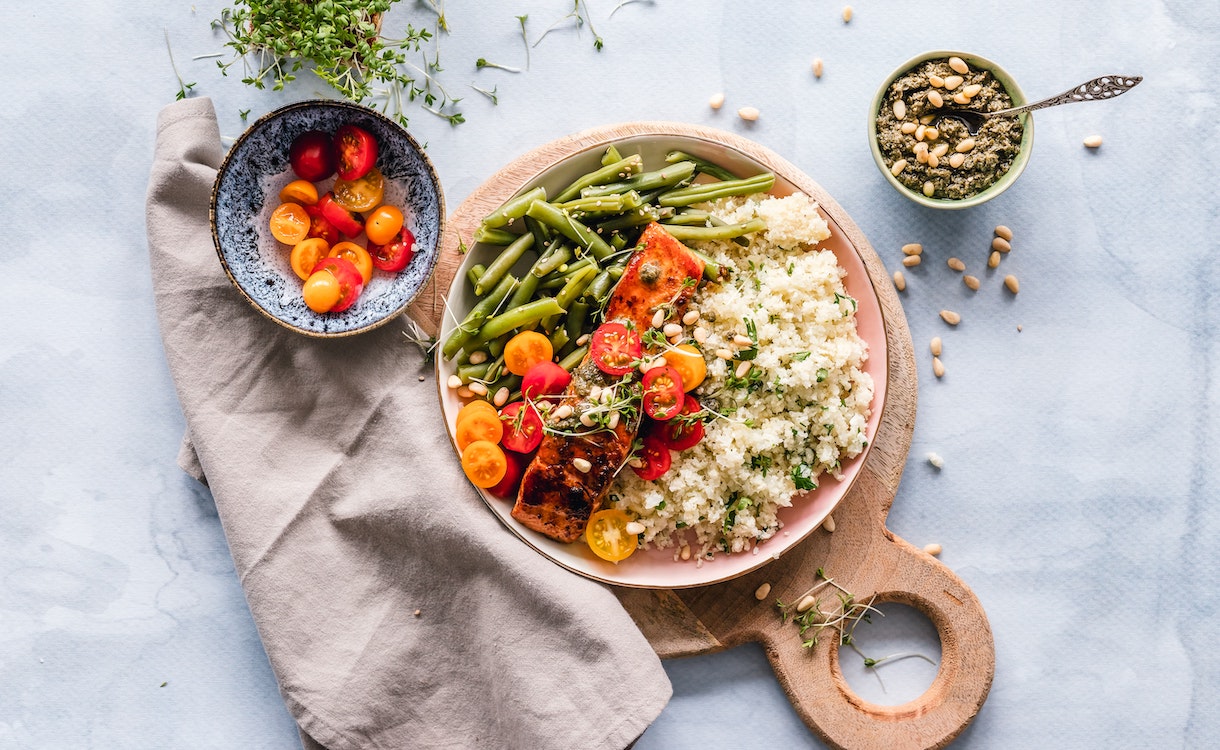 Plate with beans, rice, salmon, tomato and herbs on a wooden cutting board. Around it are a napkin, a bowl of cherry tomatoes, pine nuts and small bowl with pesto. 