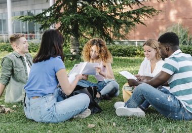 Group of young diverse men and women in casual clothes reading papers while sitting on lawn outside university building and doing homework together