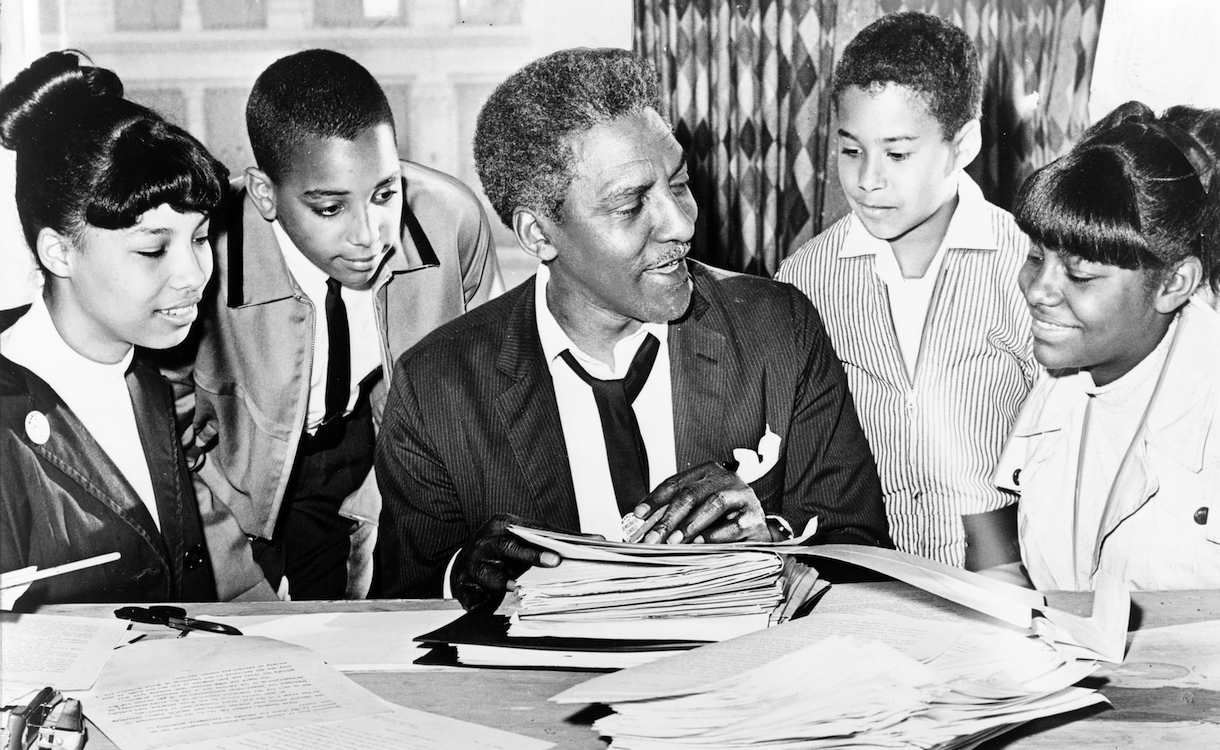 Black-and-white photo of African American civil rights leader Bayard Rustin sitting at a desk with a stack of folders in front of him, surrounded by students (left to right) Carolyn Carter, Cecil Carter, Kurt Levister, and Kathy Ross.